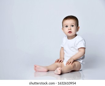 Infant Baby Boy Toddler In White Bodysuit Is Sitting Sideways On The Floor Looking Up At Us On A Gray Background With Free Text Copy Space