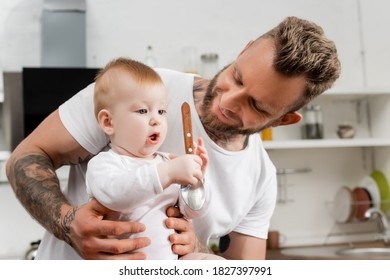 Infant Boy Holding Spoon Near Young, Bearded Father In Kitchen