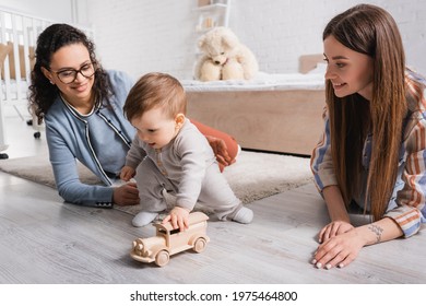 Infant Boy Playing With Wooden Toy Car Near Happy Multiethnic Women