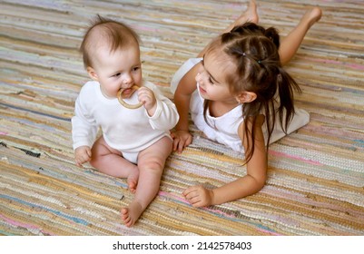 An Infant Is Gnawing A Wooden Teether Toy In The Shape Of A Ring. Drool Flows Down The Hands Of The Child. Cute Sisters Are Sitting On The Floor Of The Nursery