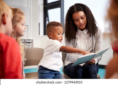 Infant school boy pointing in a book held by the female teacher, sitting with kids on chairs in the classroom, close up - Powered by Shutterstock