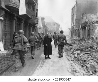 As infantrymen march through a German town, a shocked old woman stares at a the ruins. March-April 1945. Germany, World War 2.