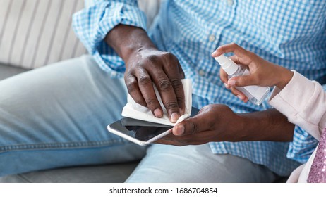 Infection Prevention Concept. African American Man And Little Child Cleaning Mobile Phone With Sanitizer, Closeup. Empty Space