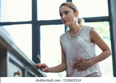 Infection Of Stomach. Low Angle Of Worried Mature Woman Touching Belly And Leaning On Surface