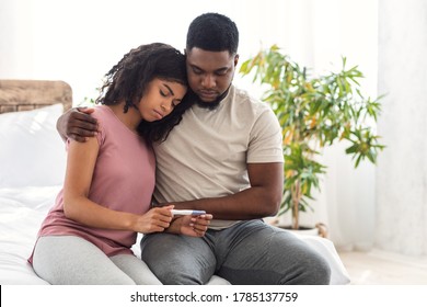 Infertility concept. African sad guy hugging his wife holding negative pregnancy test, bedroom interior, free space
