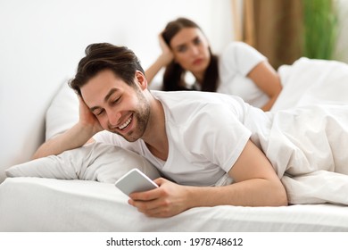 Infidelity. Cheating Husband Texting On Phone With Lover Ignoring Unhappy Jealous Wife Lying In Bed At Home. Male Cheater Having Affair. Jealousy And Unfaithfulness In Relationship. Selective Focus