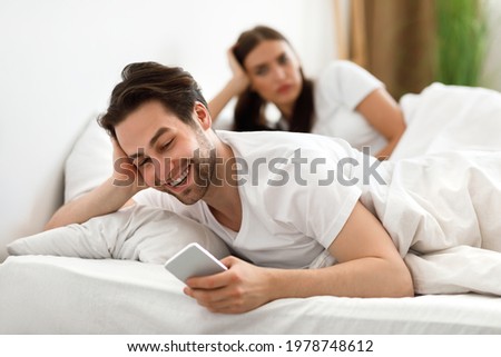 Infidelity. Cheating Husband Texting On Phone With Lover Ignoring Unhappy Jealous Wife Lying In Bed At Home. Male Cheater Having Affair. Jealousy And Unfaithfulness In Relationship. Selective Focus