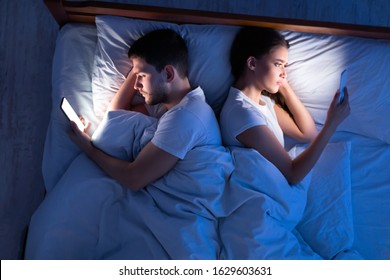 Infidelity. Cheating Spouses Using Smartphones Texting With Lovers Lying Back-To-Back In Bed At Home At Night. Top View, Low-Light