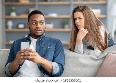 Infidelity Concept. Suspicious Young Woman Watching Her Black Boyfriend Texting On Phone, Jealous Lady Trying To Read Messages While Relaxing In Living Room Together, Suspecting Unfaithfulness