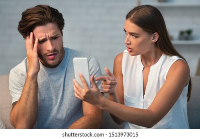 Infidelity Concept. Wife Showing Her Cheating Husband His Cellphone Demanding Explanation Sitting On Couch At Home.