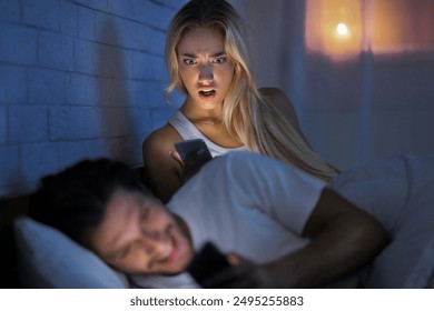 Infidelity. Jealous Girlfriend Catching Boyfriend Texting On Smartphone With Other Women Lying In Bed. Low Light, Selective Focus - Powered by Shutterstock