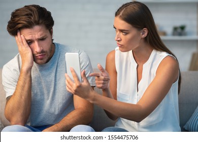Infidelity. Jealous Girlfriend Showing Her Cheating Boyfriend His Phone Demanding Explanation Sitting On Sofa Indoor