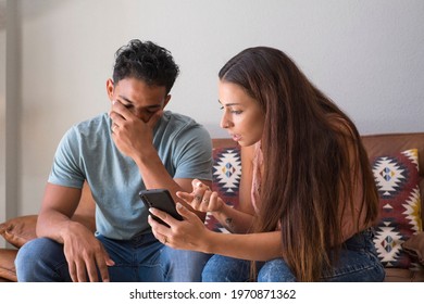 Infidelity. Jealous girlfriend Showing his Cheating boyfriend his Phone Demanding Explanation Sitting On Sofa Indoor at home. Wife caught her husband while cheating with mobile phone - divorce  people
