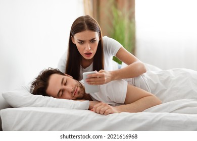 Infidelity. Shocked Wife Reading Message From Lover On Phone While Cheating Husband Sleeping In Bedroom At Home. Jealous Woman Checking Boyfriend's Cellphone Chats. Affair, Relationship Issues