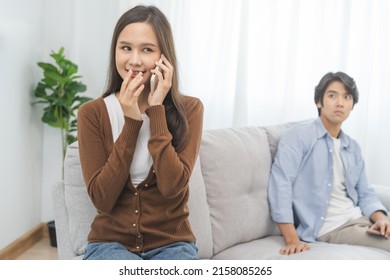 Infidelity, suspicion asian young couple love, wife jealous gossip talking mobile phone, husband spying his girlfriend while sitting on couch at home,sneaky distrust and jealousy, relationship problem