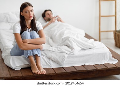 Infidelity. Unhappy Girlfriend Sitting On Bed While Cheating Husband Texting On Phone Lying In Bedroom Indoor. Sad Unloved Wife Suffering From Jealousy Suspecting Affair. Selective Focus