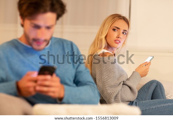 Infidelity. Wife Catching Cheating Husband Texting\
On Cellphone With Another Woman Sitting On Sofa At Home. Selective\
Focus