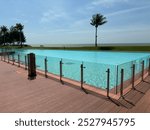 Infinity pool with modern glass railings by the ocean, surrounded by a wooden deck and palm trees