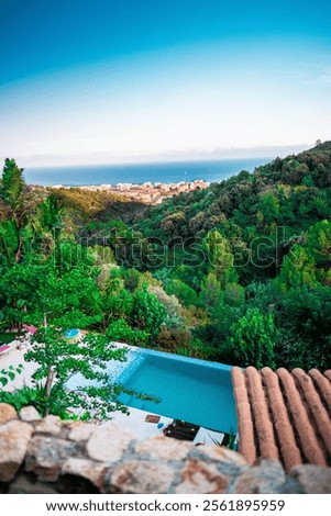 Infinity pool in a villa surrounded by green hills with a coastal town visible in the background under a bright blue sky.