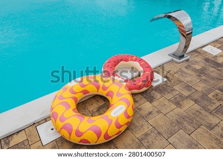 Inflatable pool floats shaped like a donut and a swirl pattern are placed next to a turquoise swimming pool featuring a sleek water fountain