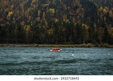 An inflatable rubber boat with rafters in red life jackets and yellow helmets are rafting on a mountain river, against the background of bright autumn mountains covered with thick fog and clouds.