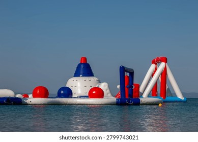 Inflatable water park structure, featuring white, blue, and red buoyant elements, floating on the clear blue surface of the sea under a bright, cloudless sky