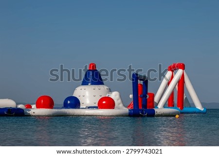 Inflatable water park structure, featuring white, blue, and red buoyant elements, floating on the clear blue surface of the sea under a bright, cloudless sky