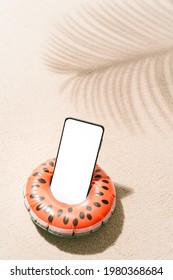 Inflatable watermelon shaped with a mobile phone inside on tropical beach sand with shadows of coconut tree leaves in summer. Travel and holiday concept background.vertical image with copy space