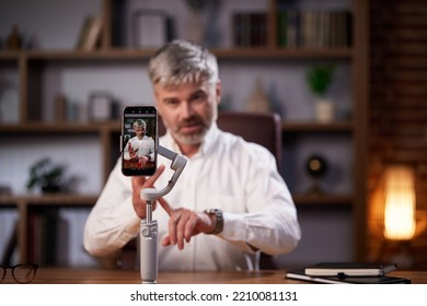 Influential, Blogger Businessman Using A Phone On A Tripod Records A Video Blog From His Home Office. Successful Man Actively Chatting Live Webinar Using Smartphone While Sitting At Workplace