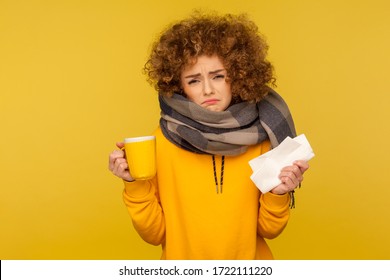 Influenza Treatment. Depressed Unhappy Flu-sick Woman With Curly Hair, Wrapped In Warm Scarf, Holding Napkin And Hot Tea, Frowning With Displeased Miserable Expression. Studio Shot, Yellow Background