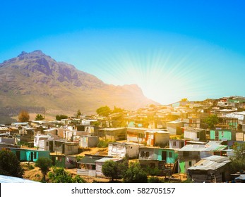 Informal Settlements, Huts Made Of Metal In The Township Or Cape Flats Of Stellenbosch, Cape Town, South Africa With Big Mountain And Blue Sky And Gold Light Background