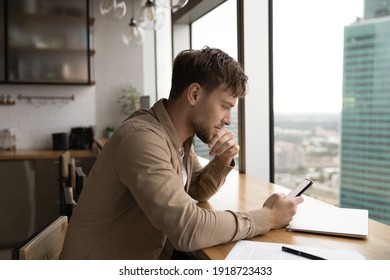 Information to think about. Pensive male look at cell ponder on job offer received by email. Young businessman sit by large window at modern kitchen, read important message on phone screen