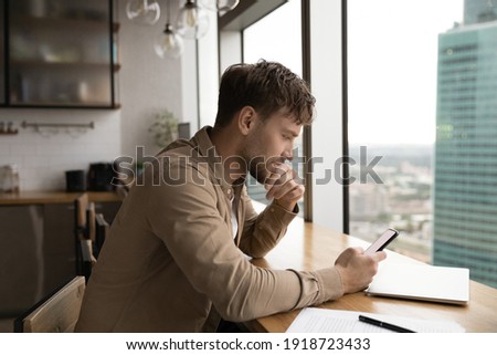 Information to think about. Pensive male look at cell ponder on job offer received by email. Young businessman sit by large window at modern kitchen, read important message on phone screen ストックフォト © 