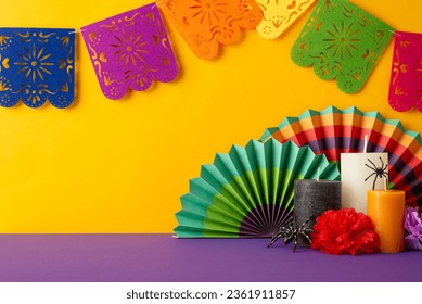 Infuse Day of the Dead vibes. Side view photo of a purple tabletop featuring vibrant fans, floral arrangements, candles, spiders, and a lively flag garland against a yellow wall, with text space