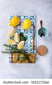 Ingedients for Moroccan preserved lemon preparation: fresh lemons, salt and rosemary on a hand painted tile with Moroccan preserved lemons jar in the bottom. Preparation Process. Top View
