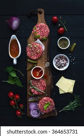 Ingredients For Cooking Burgers. Raw Ground Beef Meat Cutlets On Wooden Chopping Board, Onion, Cherry Tomatoes, Greens, Pickles, Tomato Sauce, Cheese, Herbs And Spices Over Black Background, Top View