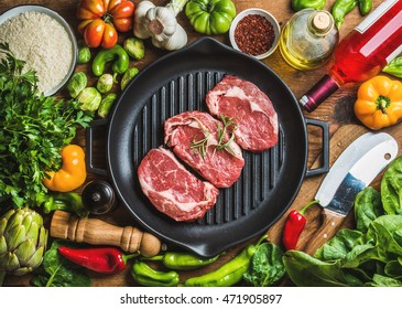 Ingredients for cooking healthy meat dinner. Raw uncooked beef steaks in cast iron grilling pan with vegetables, rice, herbs, spices and rose wine bottle over wooden background, top view - Powered by Shutterstock