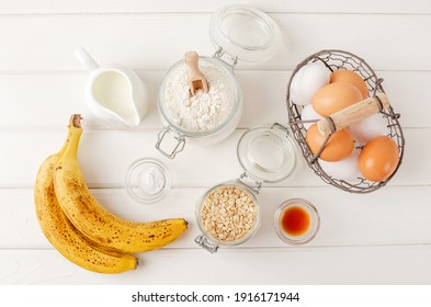 Ingredients For Cooking Oatmeal Pancakes With Bananas. Eggs, Oatmeal, Bananas, Buttermilk, Flour, Vanilla, Baking Powder On A White Wooden Background. Recipe Step By Step