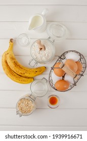 Ingredients For Cooking Oatmeal Pancakes With Bananas. Eggs, Oatmeal, Bananas, Buttermilk, Flour, Vanilla, Baking Powder On A White Wooden Background. Recipe Step By Step