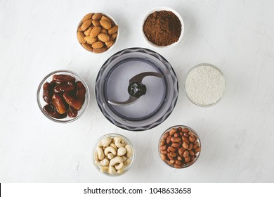 Ingredients for energy bites: nuts, dates, cocoa powder and coconut flakes with food processor on white wooden background. Step by step recipe of no bake gluten free brownie energy balls top view.