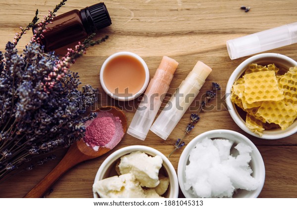 Ingredients for\
homemade lip balm: shea butter, essential oil, mineral color\
powder, beeswax, coconut oil. Homemade lip balm lipstick mixture\
with ingredients scattered\
around.