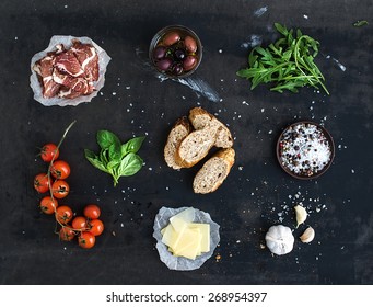Ingredients for sandwich with smoked meat, baguette, basil, arugula, olives, cherry-tomatoes, parmesan cheese, garlic and spices over black grunge background. Top view