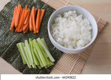 Ingredients for sushi: sliced carrots and celery, bowl with rice, nori and a bamboo mat on a work plate.