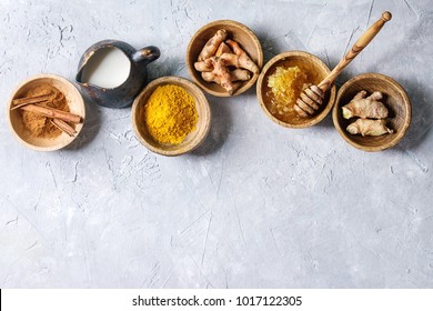 Ingredients for turmeric latte. Ground turmeric, curcuma root, cinnamon, ginger, honeycombs in wooden bowls, jug of milk over grey texture background. Top view, copy space