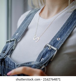 Initials Necklace Around The Neck Of A Girl With Blond Hair, Denim Salopet And White Outfit. Image For E-commerce, Online Sales, Social Media, Jewelry Sale.