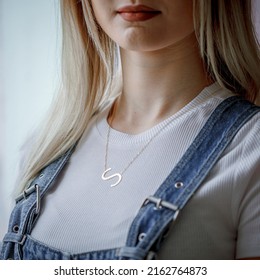 Initials Necklace Around The Neck Of A Girl With Blond Hair, Denim Salopet And White Outfit. Image For E-commerce, Online Sales, Social Media, Jewelry Sale.