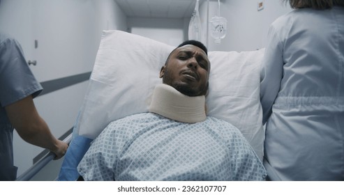 Injured African American man lies on gurney in neck brace and tries to talk with medics. Female doctor and nurse transport patient to hospital room. Medical staff at work in modern medical center. - Powered by Shutterstock