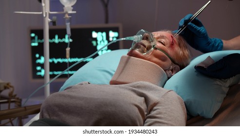 Injured Aged Woman With Neck Brace And Oxygen Mask Sleeping In Hospital Bed While Nurse Taking Glass Shards From Wound On Her Forehead. Doctor Treating Bruise Of Senior Female Patient