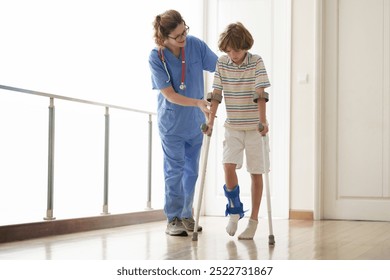 Injured child on crutches. Boy in cast on foot and leg. Little kid with sprained ancle learning to walk again. Physiotherapy in health clinic. Orthopedic trauma treatment. Doctor helping young patient - Powered by Shutterstock