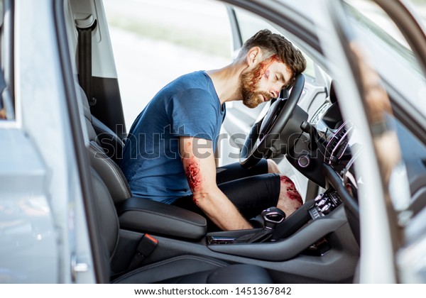 Injured man with a broken head and bleeding wounds\
sitting on the driver seat without consciousness after the road\
accident inside the car